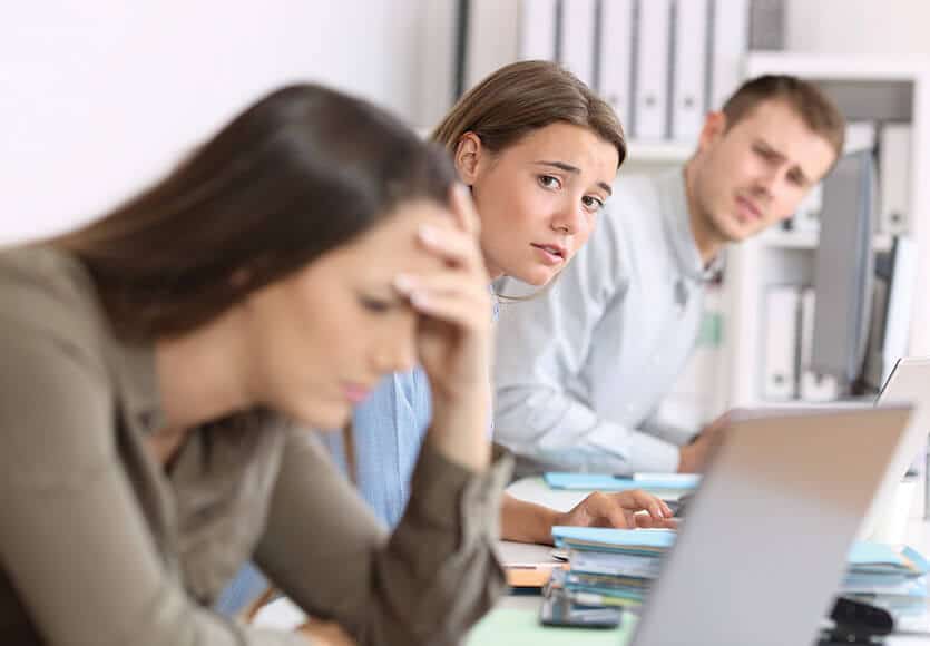 Irodai környezetben egy nő és egy férfi aggódva nézi, ahogy egy másik nő a homlokán tartja a kezét és aggódva nézi a monitorját.