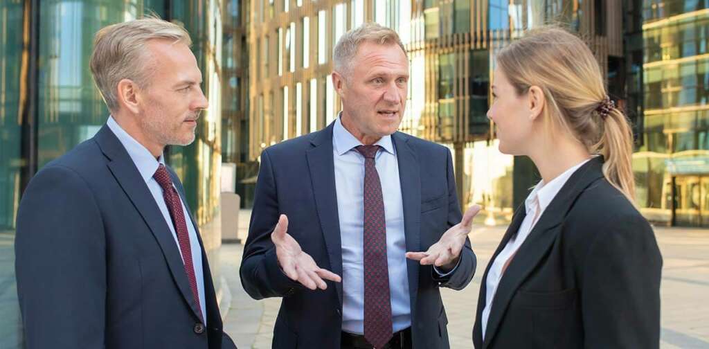 Üzleti ruházatba öltözött emberek, két férfi és egy nő beszélgetnek egy utcán. A középen álló férfi magyaráz, a másik két személy őt figyeli.